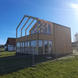 Surowa bryła dwukondygnacyjnego domu w trakcie budowy, z widoczną drewnianą konstrukcją szkieletową dachu i dużymi oknami na parterze, otoczona tymczasowym ogrodzeniem i zielonym trawnikiem.