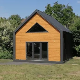 Niewielki, nowoczesny budynek z drewnianą elewacją w odcieniach brązu i czarnym dachem z blachy na tle zielonego trawnika i drzew. Budynek ma duże, przesuwne drzwi i trójkątne okno pod dachem.