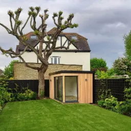 Nowoczesna, drewniana konstrukcja z przeszkloną ścianą, umieszczona w ogrodzie z równo przystrzyżonym trawnikiem, obok fantazyjnie przyciętego drzewa, na tle domu z muru pruskiego.