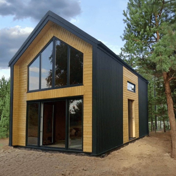 Nowoczesny, dwukondygnacyjny budynek z dużymi przeszkleniami, wykończony drewnem i czarną blachą, otoczony drzewami na piaszczystym podłożu.