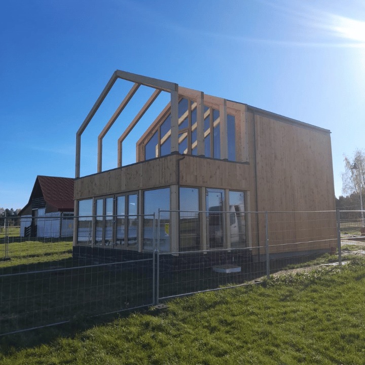 Surowa bryła dwukondygnacyjnego domu w trakcie budowy, z widoczną drewnianą konstrukcją szkieletową dachu i dużymi oknami na parterze, otoczona tymczasowym ogrodzeniem i zielonym trawnikiem.