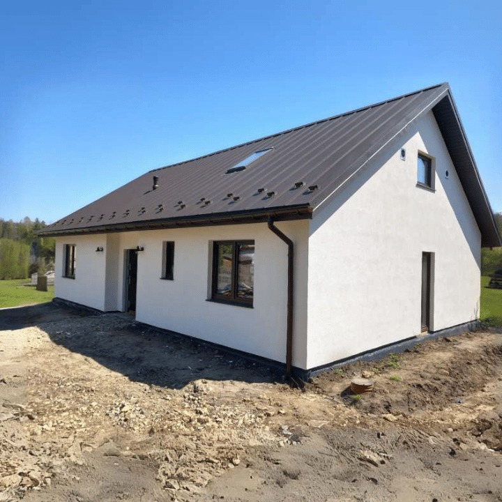 Niedawno wybudowany, parterowy dom z białymi ścianami i ciemnobrązowym dachem z blachy, widoczny z zewnątrz, na niezagospodarowanej działce z ziemią i gruzem.