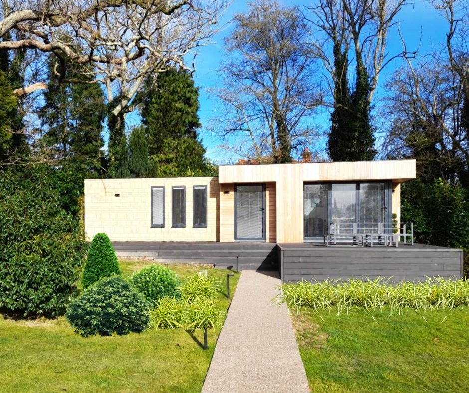Nowoczesny, parterowy budynek z jasnego drewna z płaskim dachem i dużymi oknami, otoczony zielenią, z przylegającym tarasem z desek kompozytowych i ścieżką prowadzącą do wejścia.