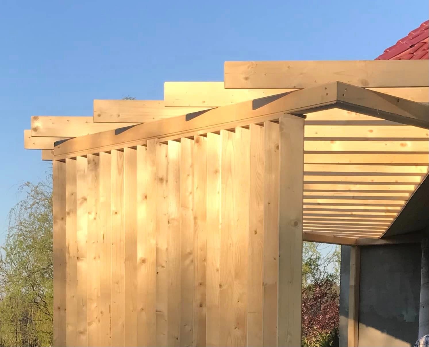 Konstrukcja drewniana w trakcie budowy, widoczna surowa ściana z pionowych desek i zadaszenie z poziomych listew, z fragmentem czerwonego dachu w tle, oświetlona popołudniowym słońcem.