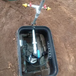 Studzienka zaworowa z pompą głębinową w czarnej obudowie, instalacja wodna z zaworami na powierzchni, otoczona ziemią.