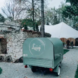 Jasnozielony mobilny bar Piaggio z logo 'Luigi Mobile Bar' na tle białego namiotu eventowego i kamiennej ruiny z dekoracyjnymi kołami od wozu.
