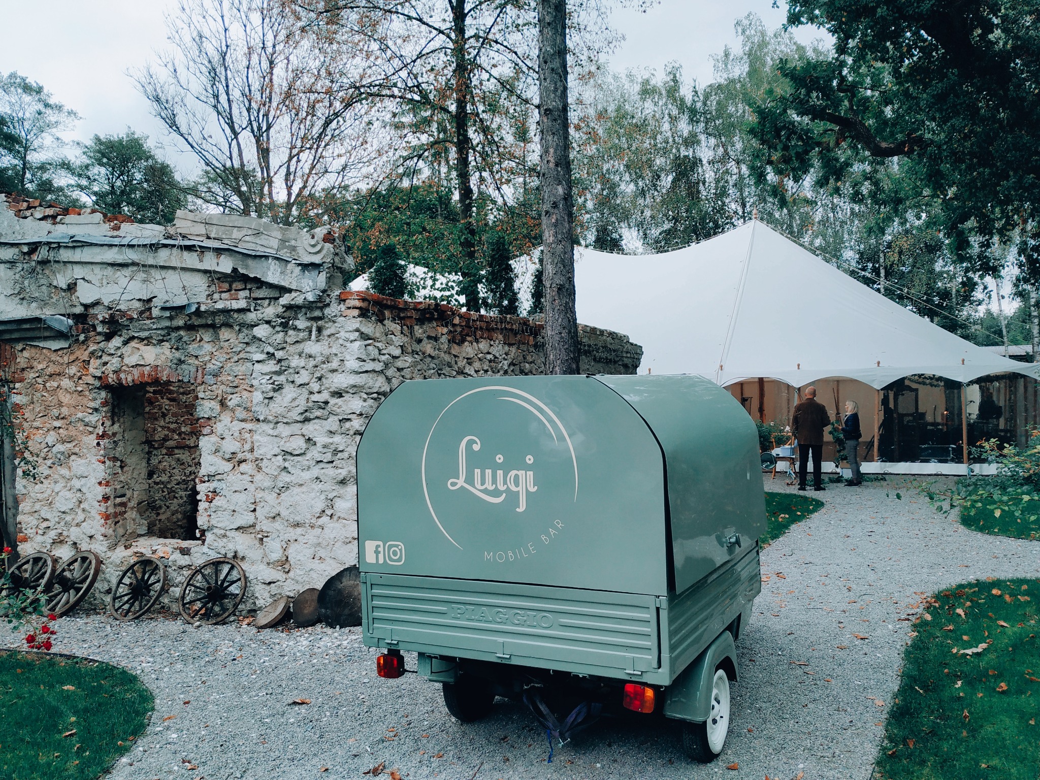 Jasnozielony mobilny bar Piaggio z logo 'Luigi Mobile Bar' na tle białego namiotu eventowego i kamiennej ruiny z dekoracyjnymi kołami od wozu.