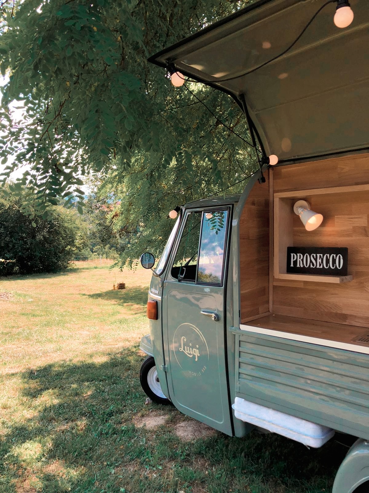 Mobilny bar w stylu retro, trójkołowy pojazd z napisem 'Luigi Mobile Bar', oświetlony girlandą żarówek, z tabliczką 'PROSECCO' w drewnianej wnęce, na tle zieleni.
