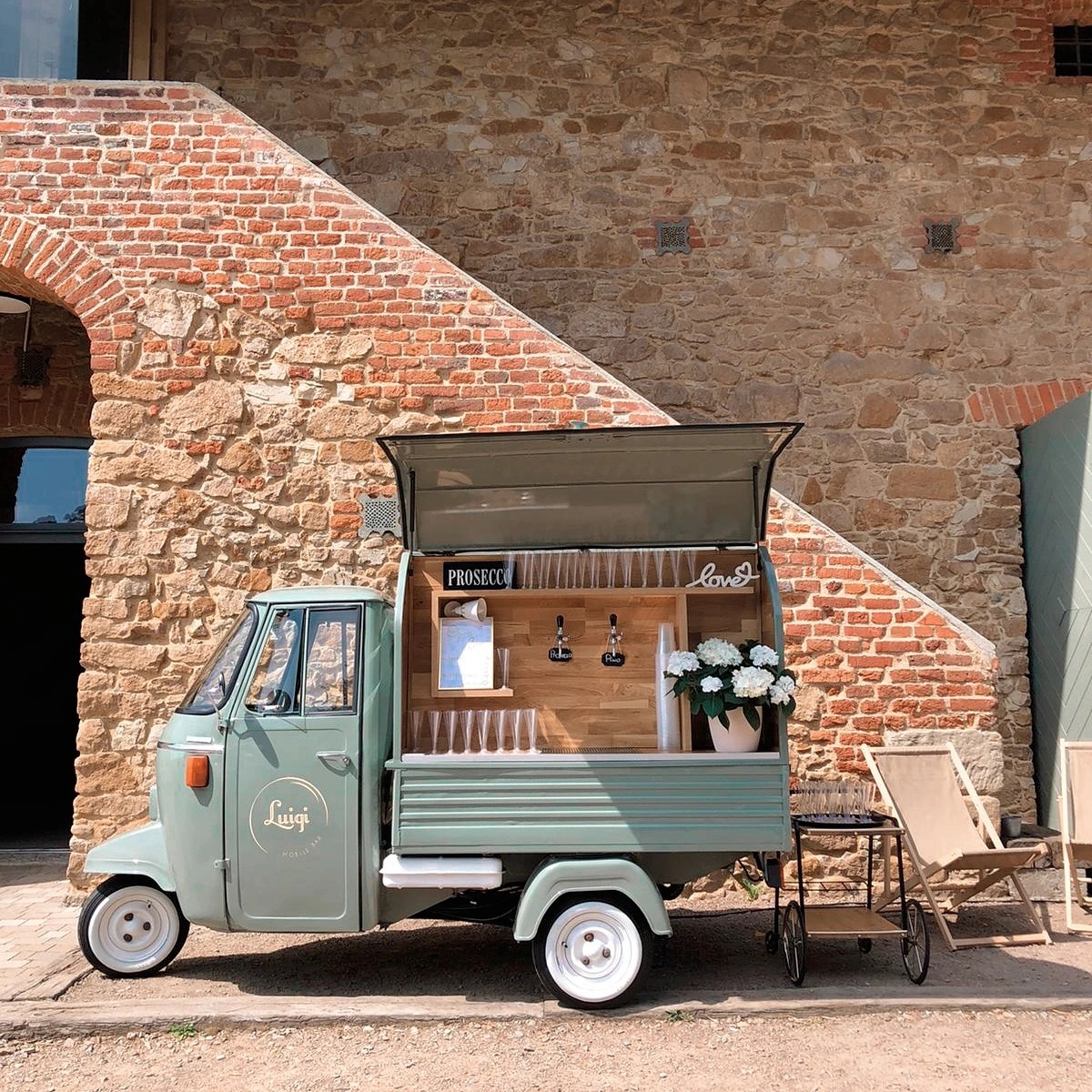 Mobilny bar Prosecco w stylu retro, umieszczony na trójkołowym pojeździe Luigi Mobile Bar, z drewnianym blatem, nalewakami do piwa, kieliszkami i kwiatami w doniczce, na tle ceglanej ściany.