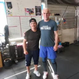 Dwóch mężczyzn pozuje w siłowni obok tablicy z planem treningowym; jeden w czapce z daszkiem i koszulce LEVEL, drugi w koszulce Reebok Posterus i kompresyjnych podkolanówkach.