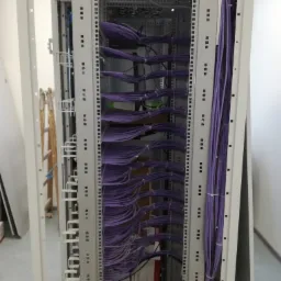 Szafa serwerowa z uporządkowanymi fioletowymi kablami, widoczne wentylatory chłodzące na górze, drabina oparta o ścianę w tle po lewej stronie.