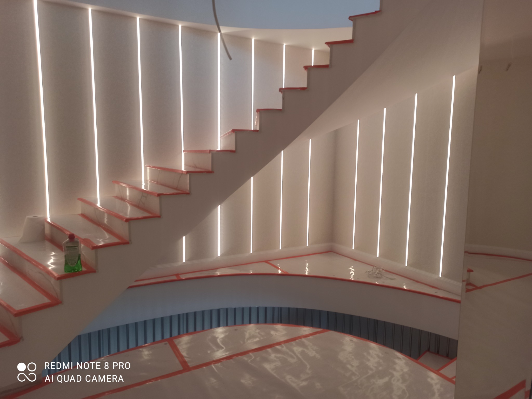 Nowoczesne schody z pionowym oświetleniem LED w ścianach, zabezpieczone folią ochronną na czas prac wykończeniowych, butelka płynu do czyszczenia na jednym ze stopni.