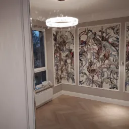 Jasne wnętrze pokoju z parkietem ułożonym w jodełkę, dekoracyjnymi panelami ściennymi z motywem ptaków i nowoczesnym okrągłym żyrandolem.