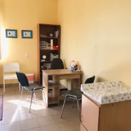 Gabinet z biurkiem, regałem i przewijakiem dla dzieci, na ścianie dyplomy w ramkach.