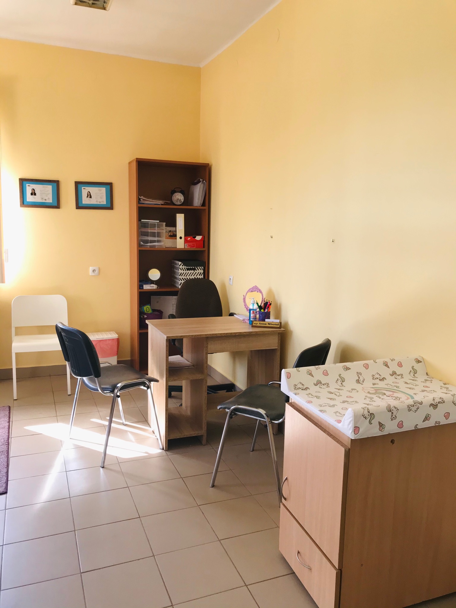 Gabinet z biurkiem, regałem i przewijakiem dla dzieci, na ścianie dyplomy w ramkach.