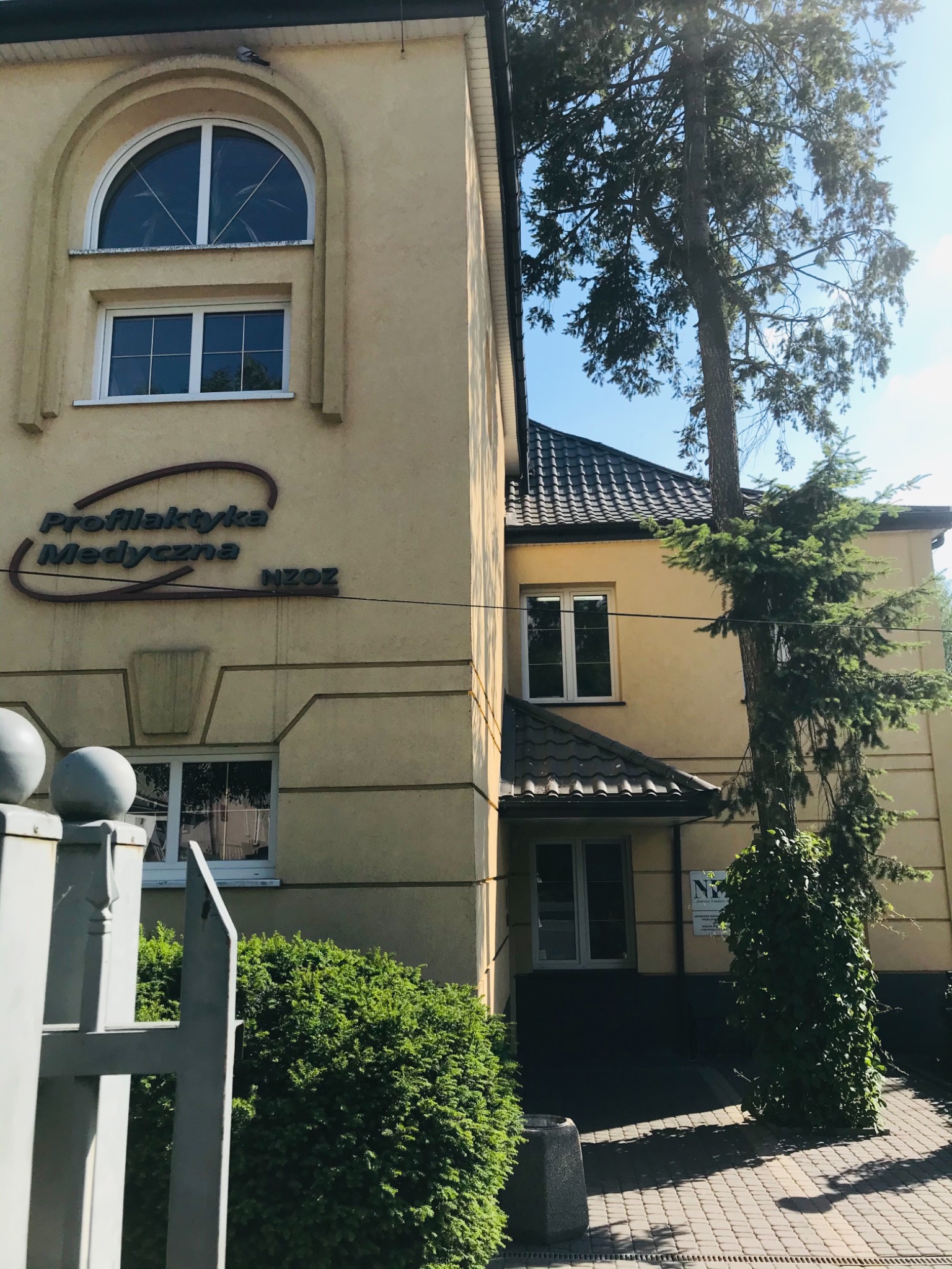 Budynek placówki medycznej z napisem 'Profilaktyka Medyczna NZOZ' i drzewem rosnącym tuż obok ściany, widok z zewnątrz.
