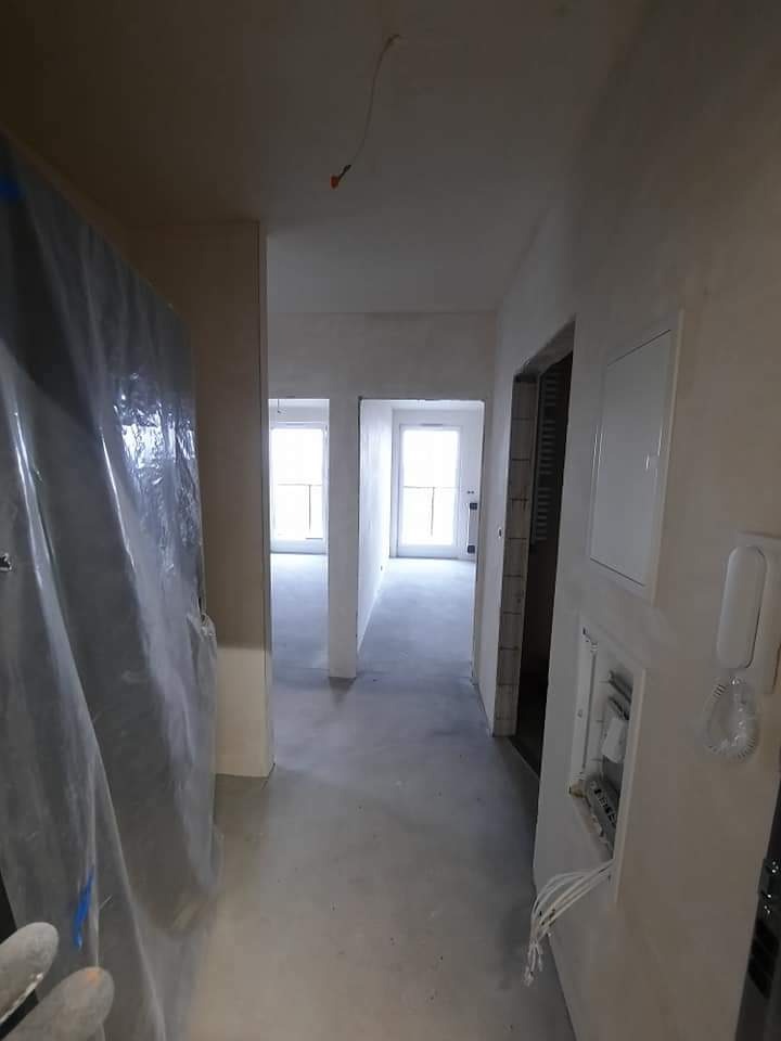 Korytarz w trakcie remontu: świeżo położone tynki maszynowe, zabezpieczona folią ściana, otwarty panel elektryczny z wystającymi kablami, widok na okna w oddali.