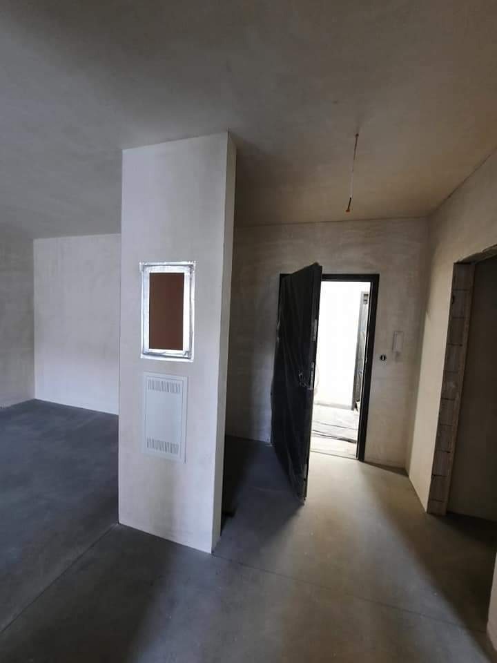 Wnętrze w stanie deweloperskim po tynkowaniu, widoczne gładkie ściany i sufit, zabezpieczone drzwi folią, oraz skrzynka instalacyjna na ścianie.