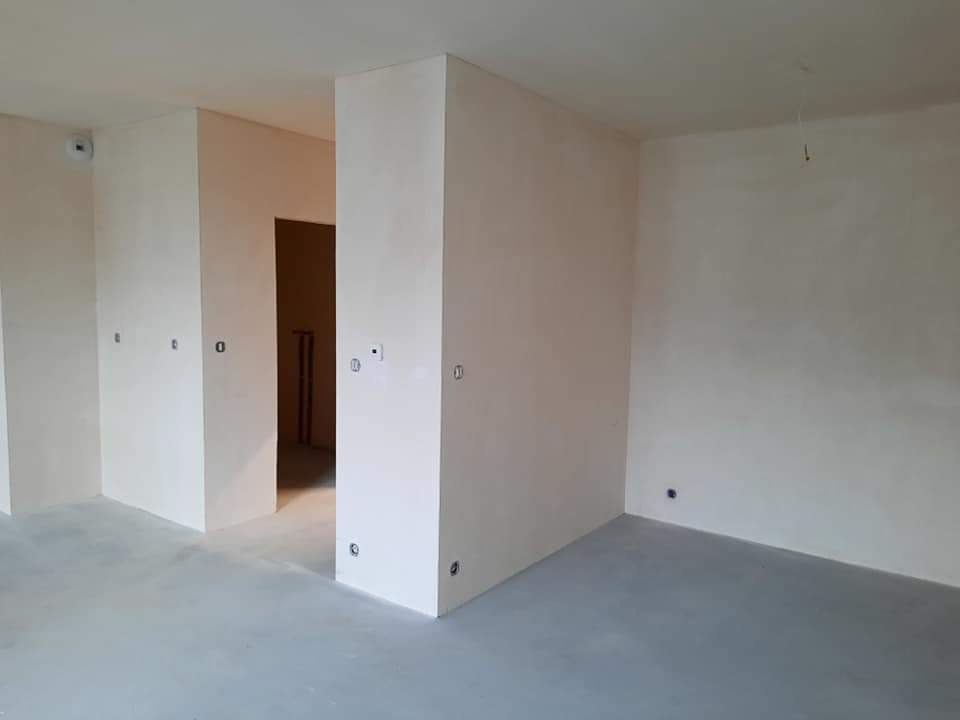 Wnętrze w trakcie prac wykończeniowych, ściany pokryte świeżym tynkiem, widoczne puszki elektryczne, szara wylewka na podłodze, surowy stan przed malowaniem.