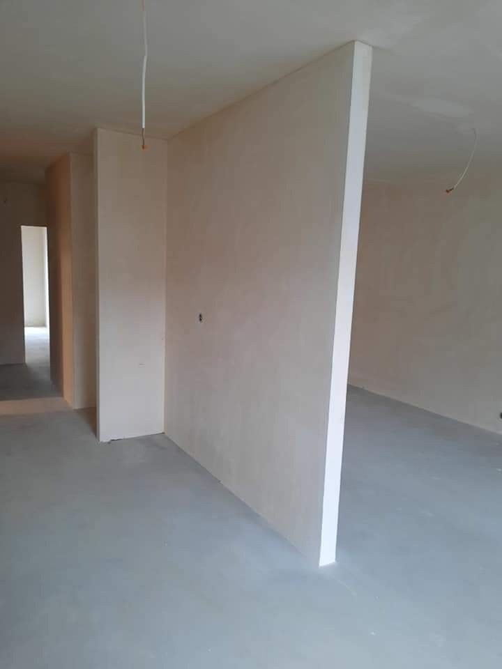 Surowe wnętrze w trakcie prac wykończeniowych: ściany pokryte świeżym, jasnym tynkiem, widoczne kable wystające z sufitu, szara wylewka podłogowa, surowy stan deweloperski.