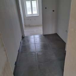 Firma usługowa Arniko - Ułożone, ciemnoszare płytki podłogowe w pomieszczeniu w trakcie remontu, z widocznymi niezagruntowanymi ścianami i fragmentem okna w tle.