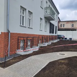Elewacja budynku po remoncie z nową kostką brukową i świeżo nawiezioną ziemią. Widoczne okna piwniczne z kratami, balkon i szara elewacja budynku.