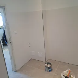 Świeżo wykafelkowana, jasna łazienka z widocznymi puszkami elektrycznymi i otwartymi drzwiami. Na podłodze stoją dwa wiadra z materiałami budowlanymi.