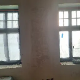 Ściana w trakcie gładzenia z dwoma oknami zabezpieczonymi folią, widoczne niedoskonałości i nierówności powierzchni, instalacja elektryczna wystająca ze ściany.