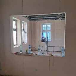 Montaż płyt kartonowo-gipsowych w pomieszczeniu z widocznymi oknami i rusztowaniem, butelki z wodą na parapecie okiennym, widoczne kable i instalacje pod sufitem.