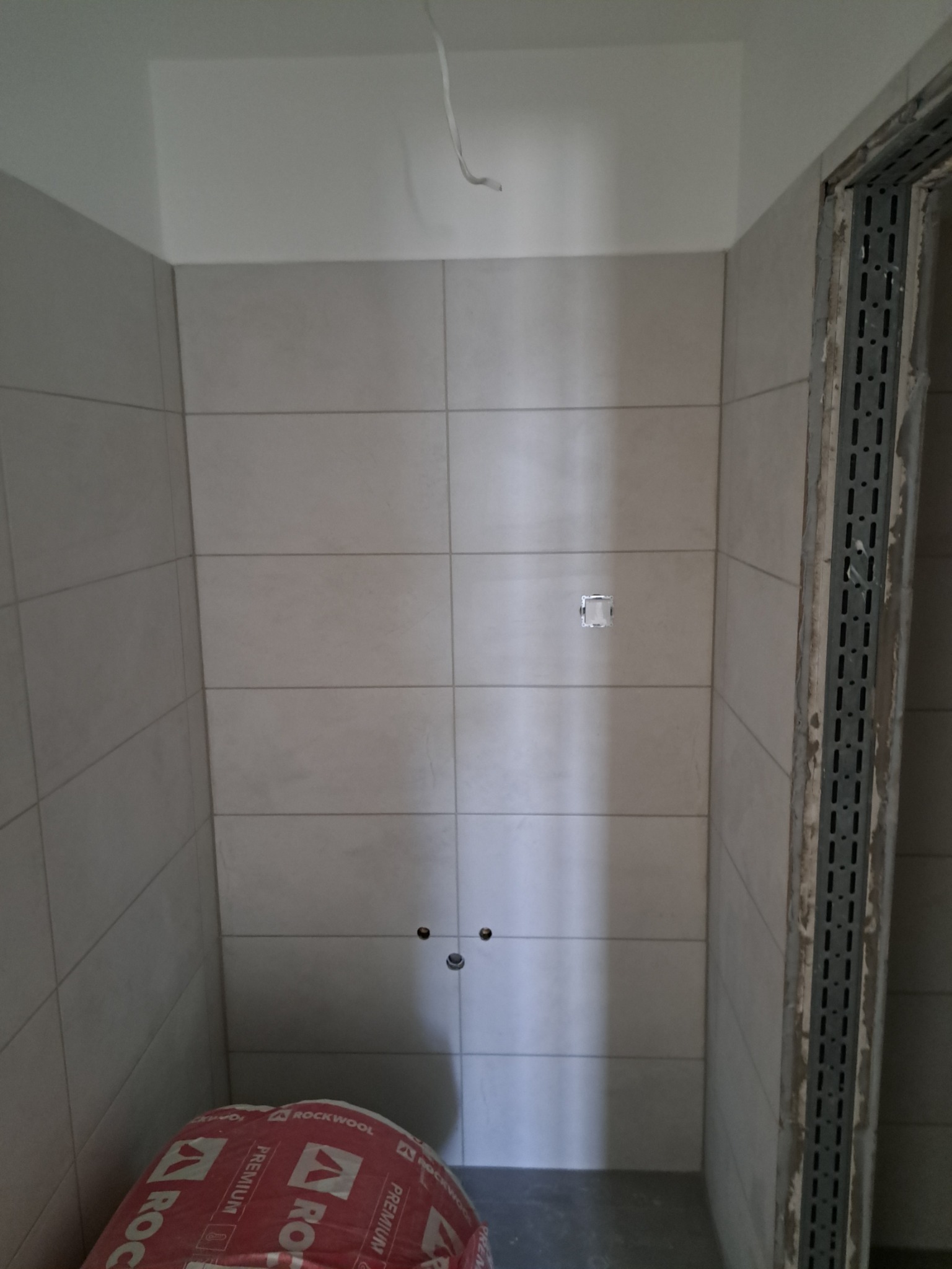 Wnętrze łazienki w trakcie prac: jasne płytki ceramiczne na ścianach, widoczne przyłącza wodne, puszka elektryczna, profile montażowe przy drzwiach oraz opakowanie wełny mineralnej Rockwool...