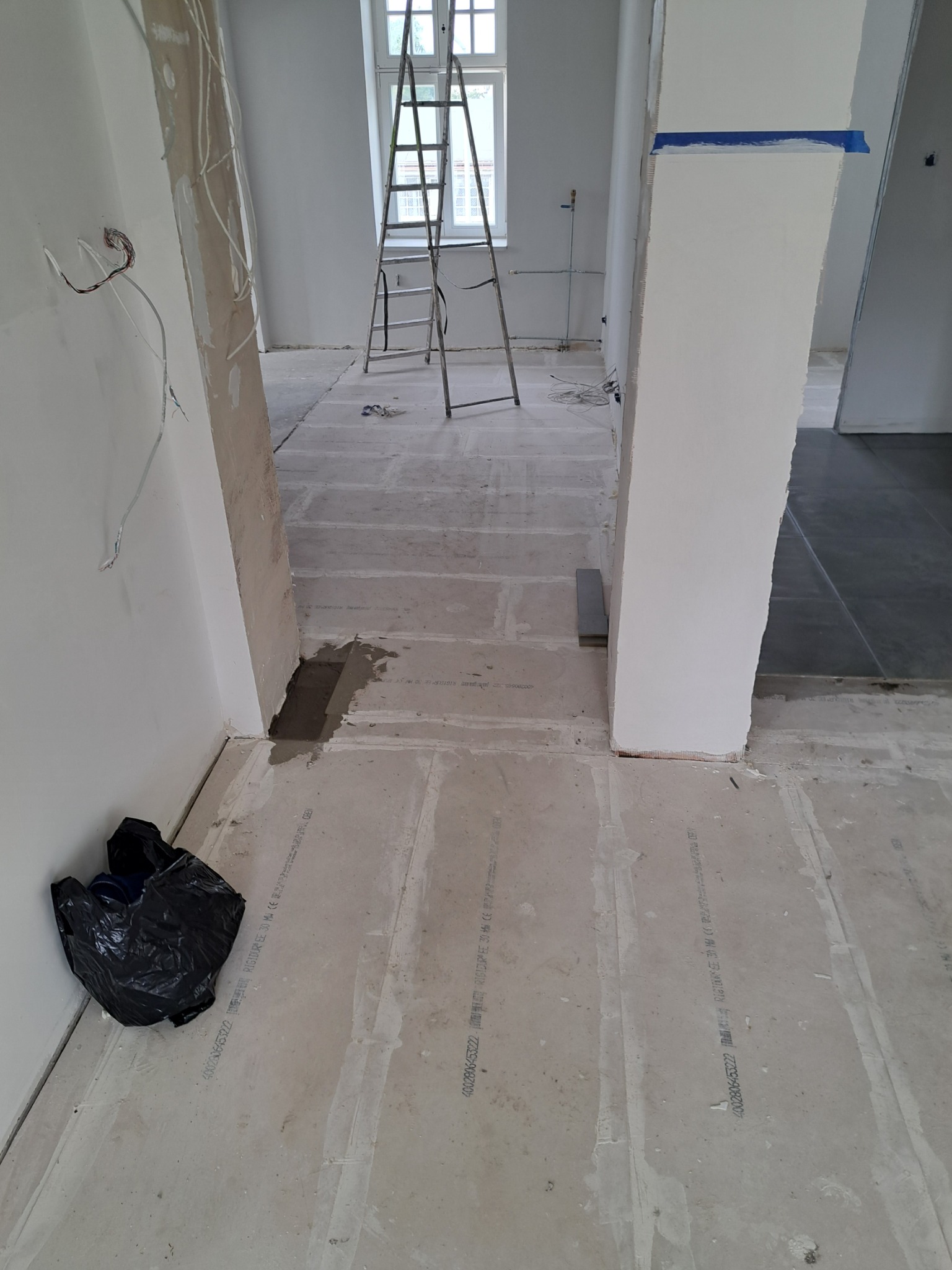 Przygotowanie podłoża z płyt RIGIDUR na podłodze, widoczne kable elektryczne wystające ze ściany, drabina aluminiowa w tle, zabezpieczenie taśmą malarską na filarze.