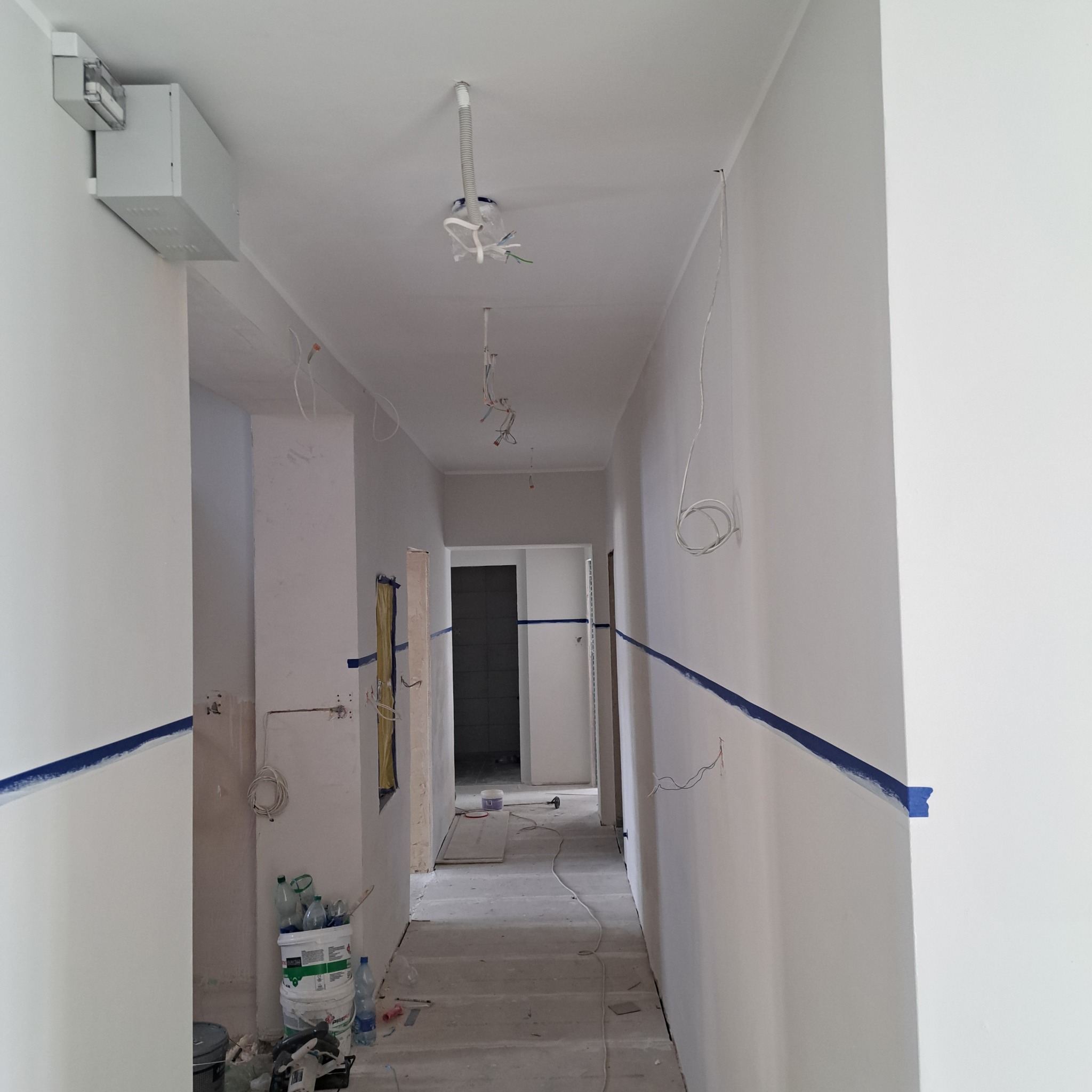 Korytarz w trakcie odświeżania ścian, z widocznymi przewodami elektrycznymi, taśmą malarską wyznaczającą linię oraz pojemnikami z farbą w tle.