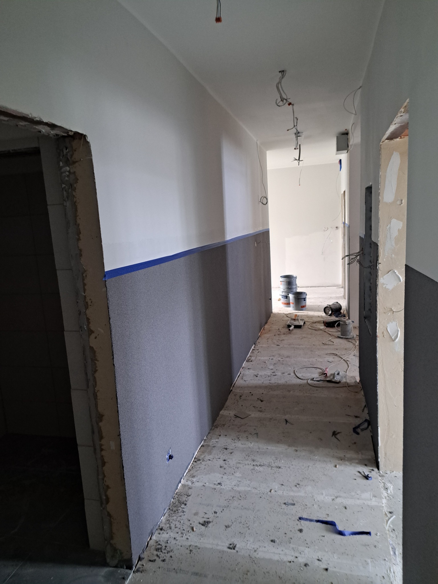 Korytarz w trakcie prac wykończeniowych w Grudziądzu: ściany częściowo pomalowane na szaro i biało, widoczne puszki z farbą, narzędzia malarskie i niezabezpieczone kable zwisające z sufitu.