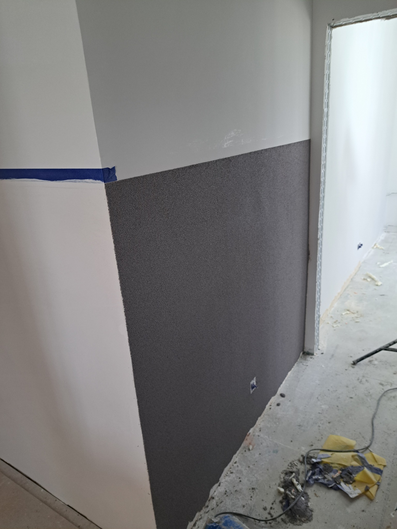 Dolna część ściany pokryta szarą, chropowatą strukturą, oddzielona niebieską taśmą malarską od górnej, białej części; widoczny fragment drzwi i podłoga w trakcie prac.