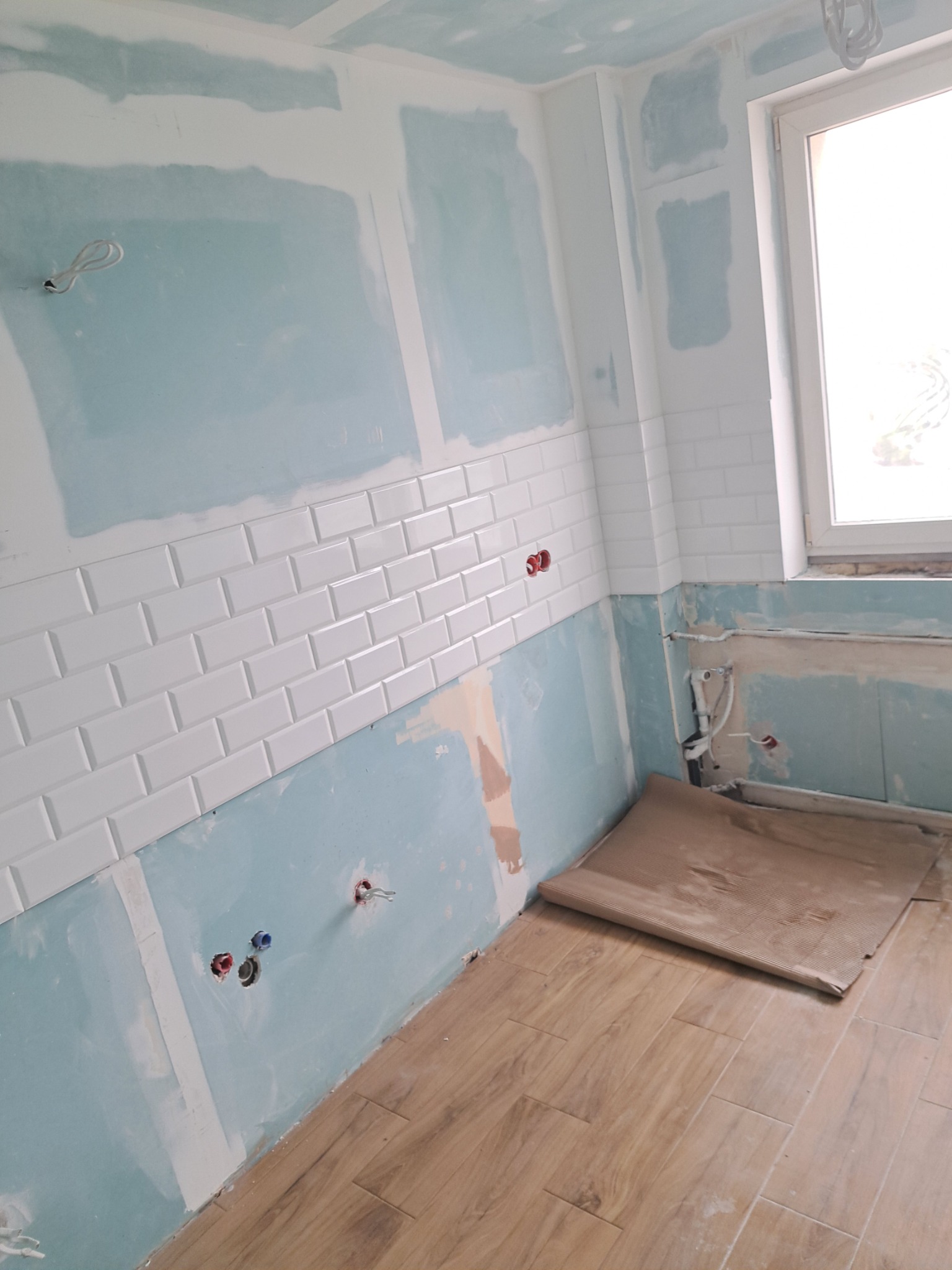 Wnętrze łazienki w trakcie remontu: położone białe płytki typu metro na ścianie, częściowo zagruntowane ściany z widocznymi instalacjami elektrycznymi i hydraulicznymi, zabezpieczona podłoga z paneli.