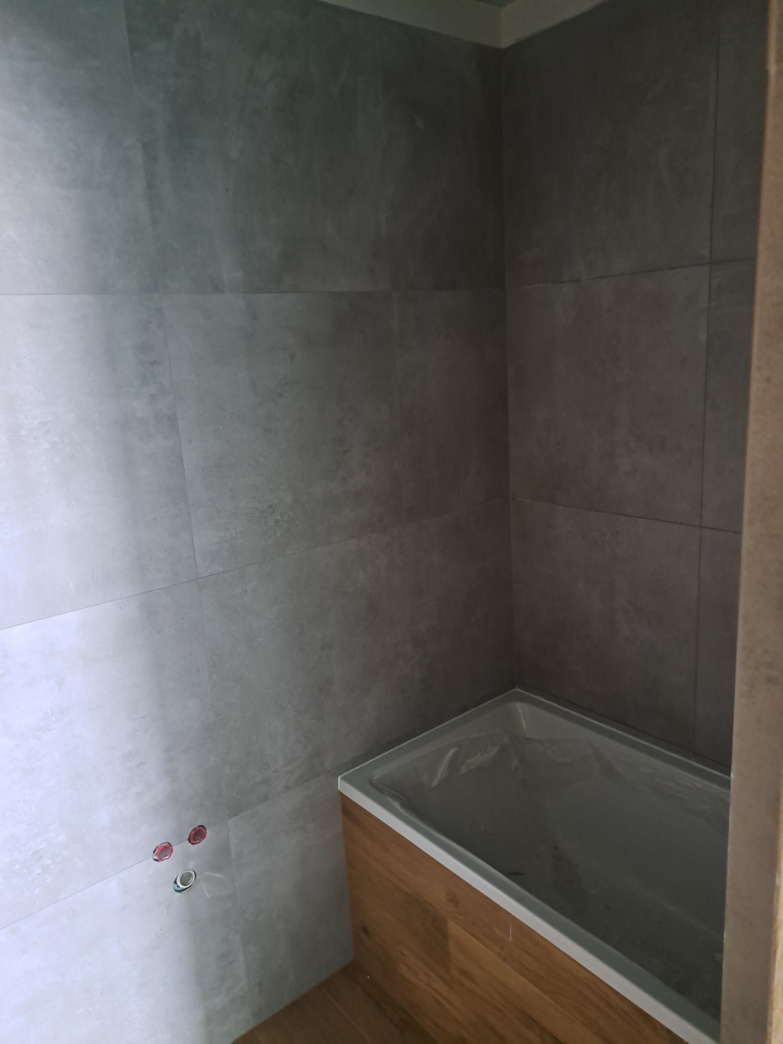 Wykończona łazienka w Grudziądzu z szarymi płytkami imitującymi kamień na ścianach i obudową wanny w kolorze drewna, widoczne przygotowane przyłącza wodne.