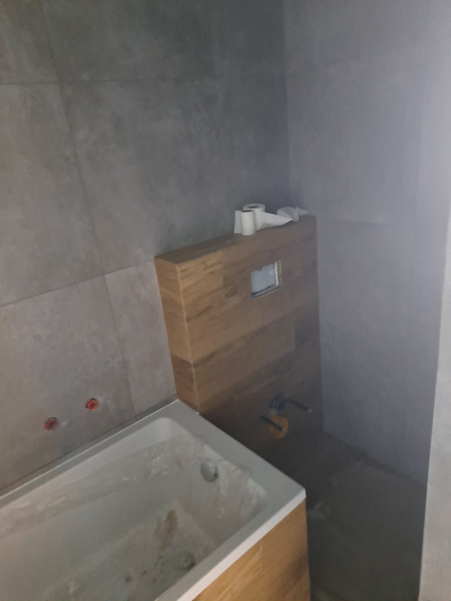 Szara łazienka w trakcie remontu: obudowa WC z drewnopodobnych paneli, biała wanna, szare płytki na ścianach, widoczne instalacje.