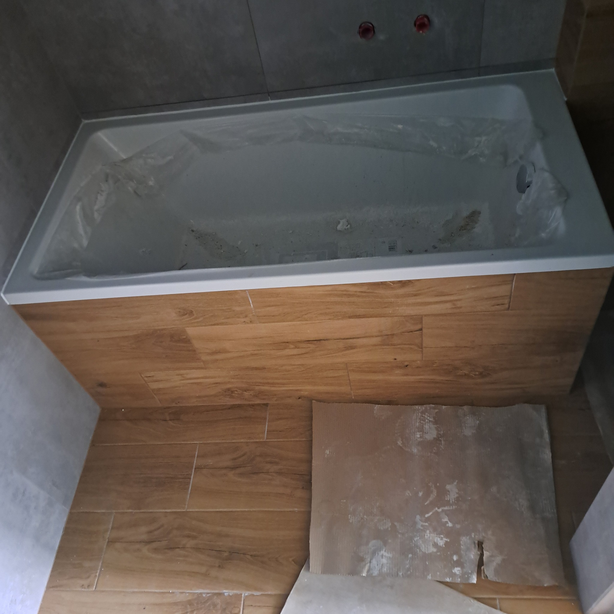 Biała wanna zabezpieczona folią, obudowana panelami imitującymi drewno, szare płytki na ścianach, karton na podłodze w trakcie prac wykończeniowych.