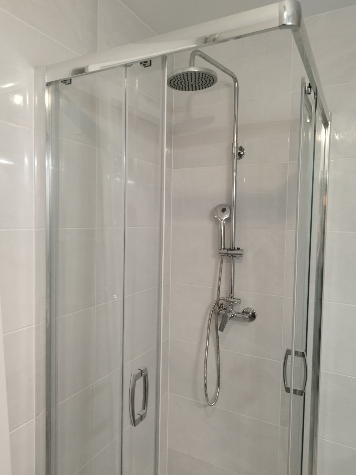 Kabina prysznicowa z przesuwnymi drzwiami, chromowaną armaturą i deszczownicą, zamontowana na ścianie wyłożonej jasnymi, kwadratowymi płytkami.