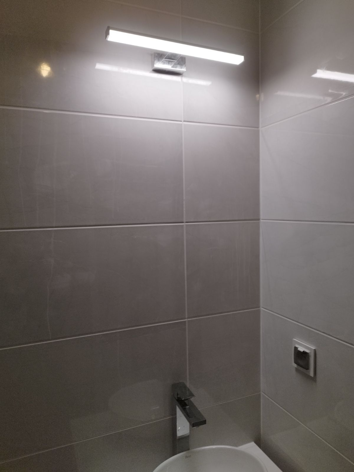 Fragment łazienki z szarymi, błyszczącymi płytkami, chromowaną baterią umywalkową i podłużnym oświetleniem LED nad umywalką. Widoczny włącznik elektryczny na ścianie.