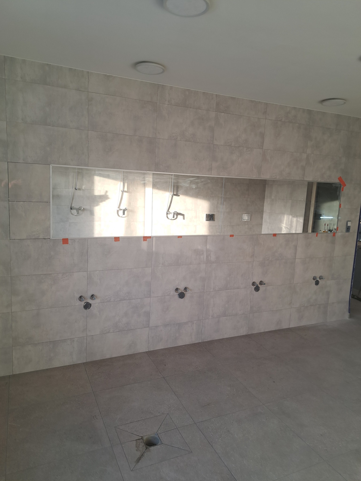 Wykończona ściana z szarych płytek ceramicznych w nowej łazience publicznej, z zamontowanym lustrem i przygotowanymi miejscami na baterie umywalkowe, widoczny odpływ podłogowy.