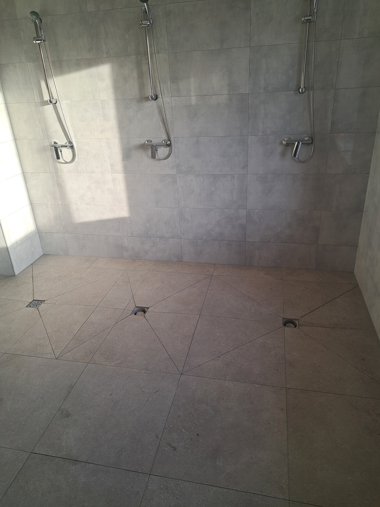 Nowoczesna kabina prysznicowa z szarymi płytkami, trzema zestawami natryskowymi i geometrycznym wzorem na podłodze z odpływami liniowymi.