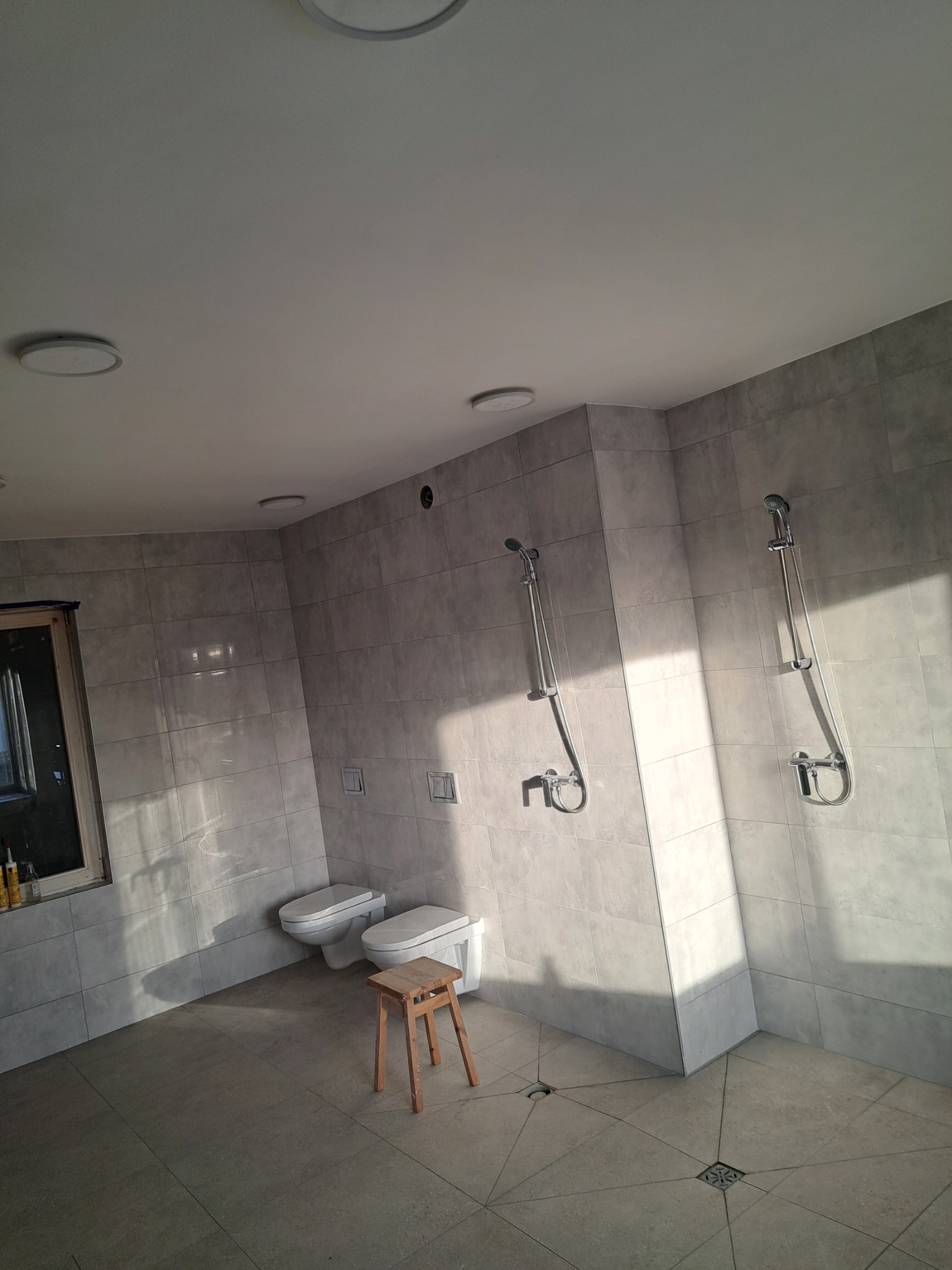Jasna łazienka z dwoma toaletami, dwoma prysznicami i drewnianym stołkiem pośrodku, wykończona szarymi płytkami.