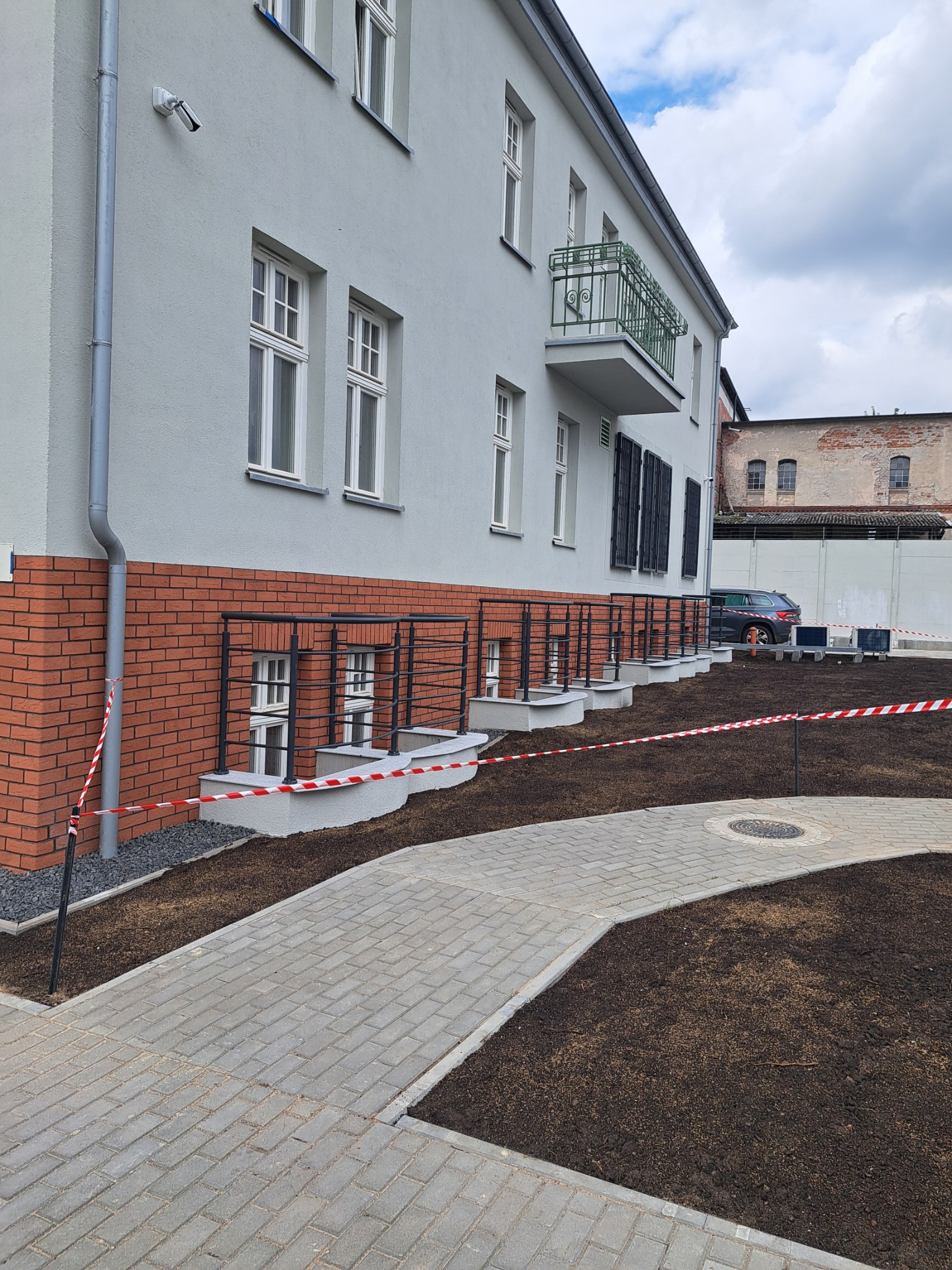 Elewacja budynku po remoncie z nową kostką brukową i świeżo nawiezioną ziemią. Widoczne okna piwniczne z kratami, balkon i szara elewacja budynku.