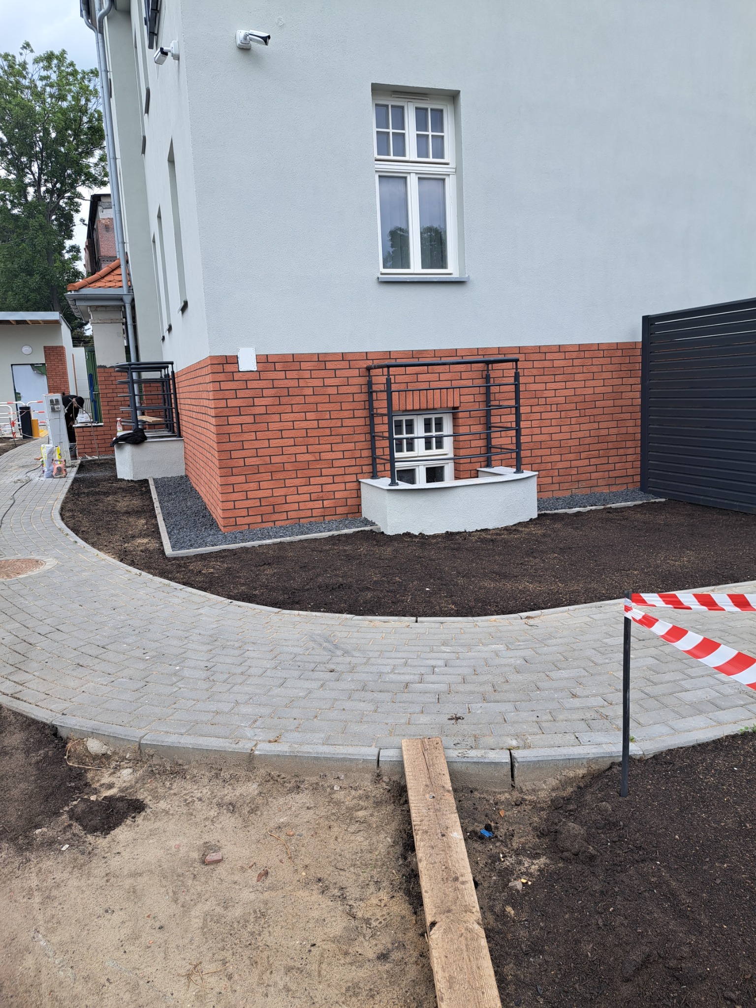 Budynek z elewacją z cegły klinkierowej, okno piwniczne z kratą, obok ułożona kostka brukowa, świeżo posadzona ziemia. Prace wykończeniowe na zewnątrz budynku.