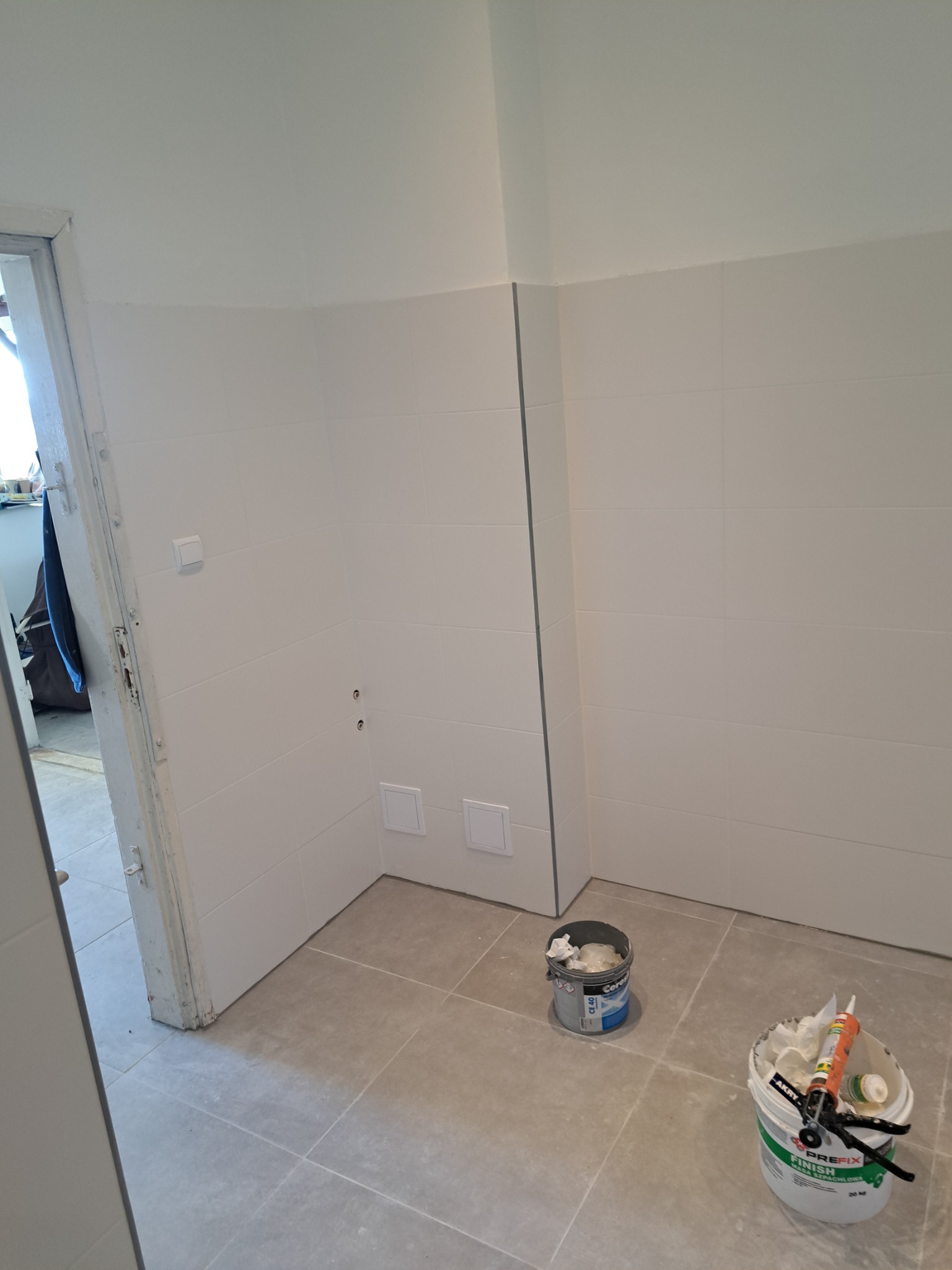 Świeżo wykafelkowana, jasna łazienka z widocznymi puszkami elektrycznymi i otwartymi drzwiami. Na podłodze stoją dwa wiadra z materiałami budowlanymi.