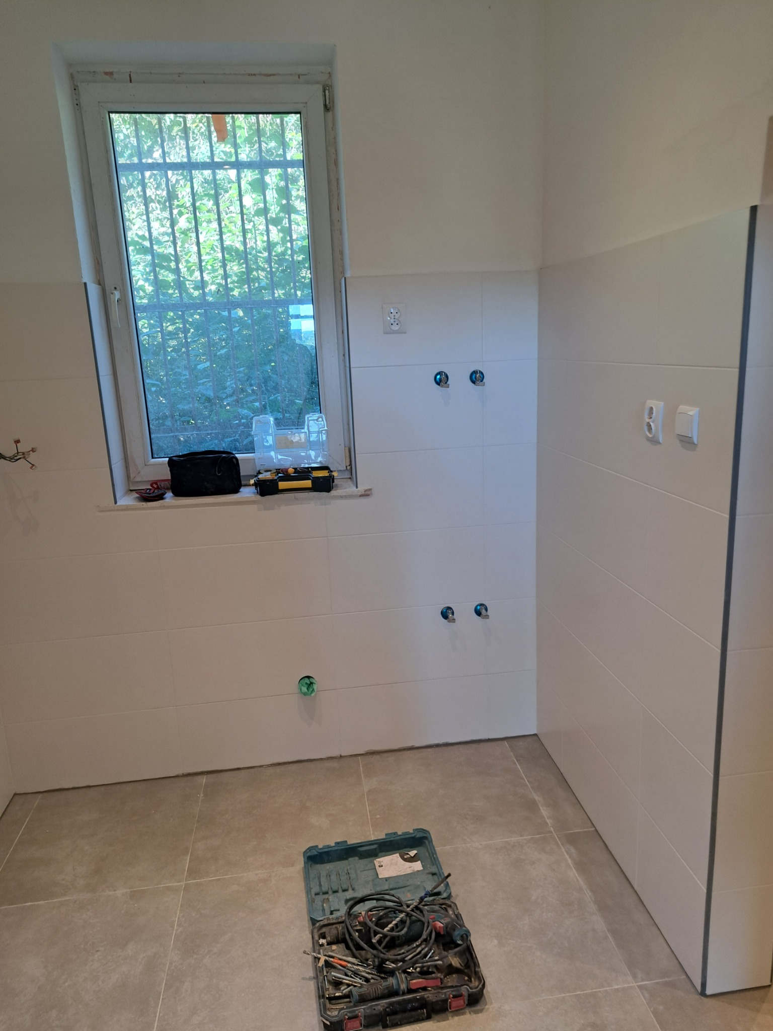 Wnętrze łazienki w trakcie remontu: białe płytki na ścianach, szare płytki na podłodze, widoczne instalacje wodne i elektryczne, skrzynka z narzędziami na podłodze.