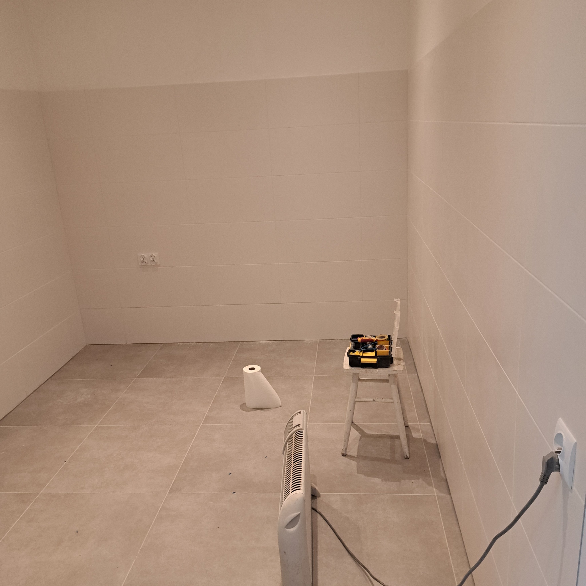 Wykończona łazienka z jasnymi płytkami, grzejnikiem i krzesłem z narzędziami. Widoczne gniazdko elektryczne i rolka papieru. Ujęcie z bliska, wnętrze po remoncie.