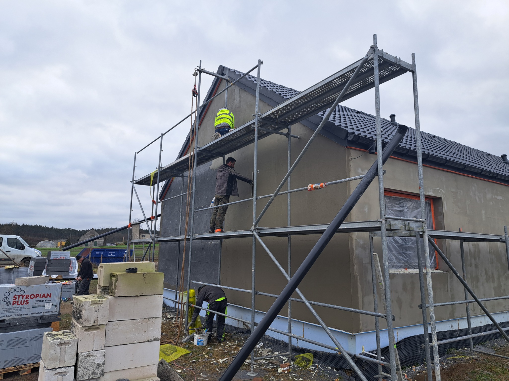 Praca na rusztowaniu przy ocieplaniu domu jednorodzinnego. Widoczne bloki styropianu, zaprawa i pracownicy w trakcie nakładania tynku. Dom z szarą dachówką.