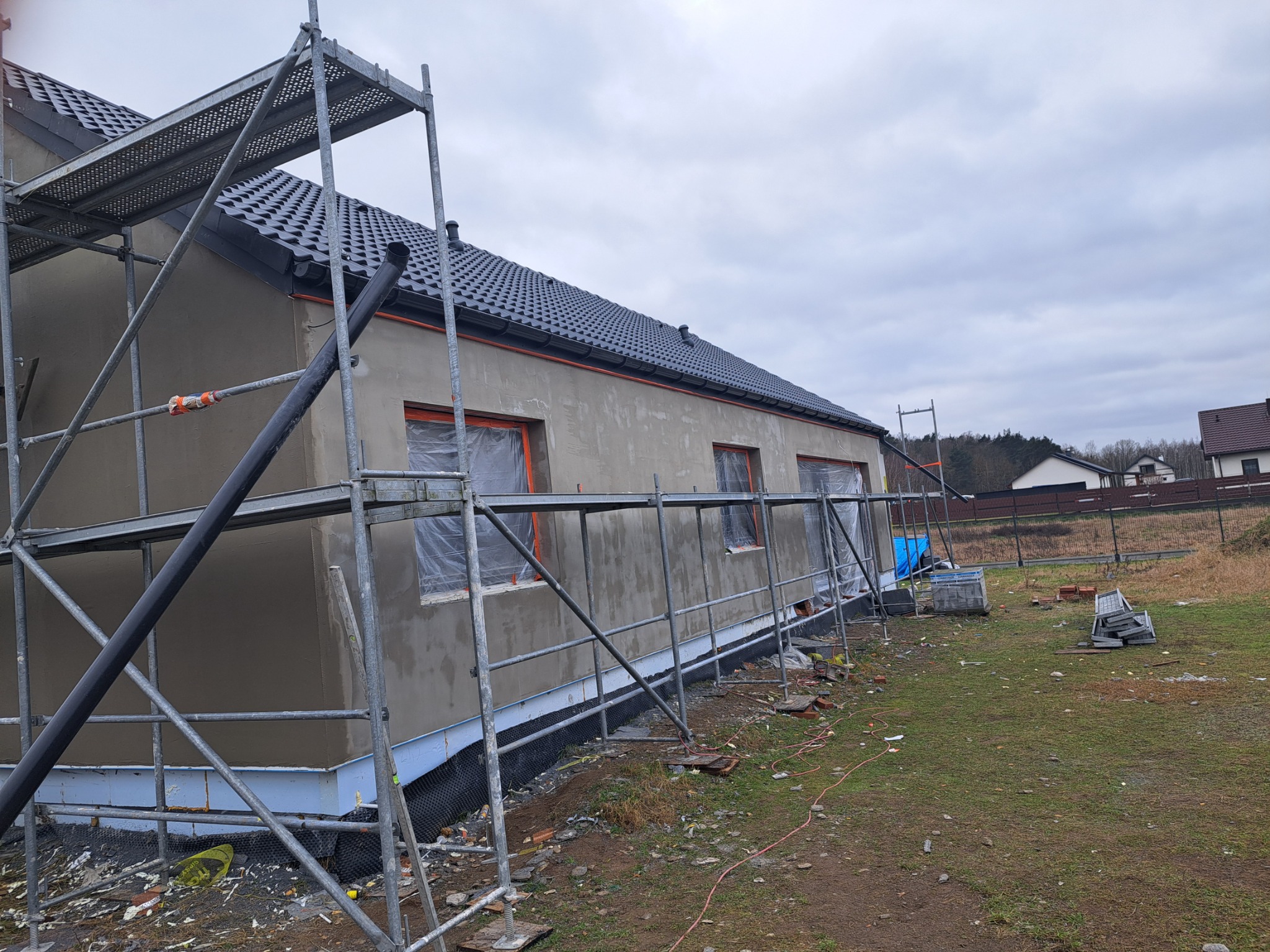 Dom jednorodzinny w trakcie prac elewacyjnych, widoczne rusztowania, okna zabezpieczone folią, świeżo nałożony tynk. Pochmurne niebo w tle.